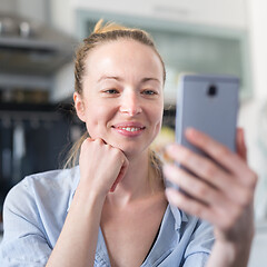 Image showing Young smiling cheerful pleased woman indoors at home kitchen using social media apps on mobile phone for chatting and stying connected with her loved ones. Stay at home, social distancing lifestyle.