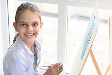 Image showing Happy teenager girl draws on an easel by the window and looked into the frame with a smile