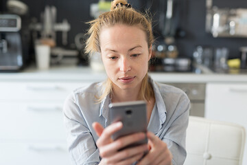 Image showing Worried woman indoors at home kitchen using social media apps on phone for video chatting and stying connected with her loved ones. Stay at home, social distancing lifestyle