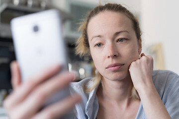 Image showing Worried woman indoors at home kitchen using social media apps on phone for video chatting and stying connected with her loved ones. Stay at home, social distancing lifestyle