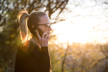 Image showing Backlit rear view of young woman talking on cell phone outdoors in park at sunset. Girl holding mobile phone, using digital device, looking at setting sun