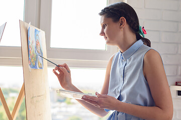 Image showing Girl draws with brushes and watercolors on an easel, sitting at home by the window