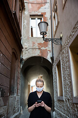 Image showing COVID-19 pandemic coronavirus. Casual caucasian woman at medieval city street using mobile phone, wearing protective face mask against spreading of coronavirus and disease transmission