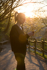 Image showing Backlit rear view of young woman talking on cell phone outdoors in park at sunset. Girl holding mobile phone, using digital device, looking at setting sun