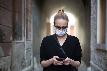 Image showing COVID-19 pandemic coronavirus. Casual caucasian woman at medieval city street using mobile phone, wearing protective face mask against spreading of coronavirus and disease transmission