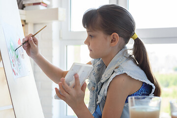 Image showing Girl diligently draws a picture on an easel in front of a large window