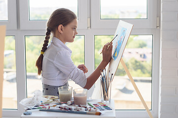 Image showing Young artist paints watercolor paints on an easel against the background of a large window