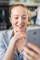 Image showing Young smiling cheerful pleased woman indoors at home kitchen using social media apps on mobile phone for chatting and stying connected with her loved ones. Stay at home, social distancing lifestyle.