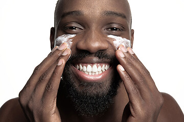 Image showing Young african-american guy applying face cream under his eyes on white background