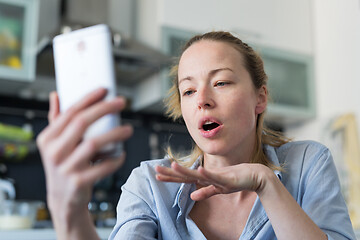 Image showing Young pleased woman indoors at home kitchen using social media apps on mobile phone for chatting and stying connected with her loved ones. Stay at home, social distancing lifestyle.