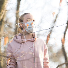 Image showing Portrait of caucasian sporty woman wearing medical protection face mask while walking in park, relaxing and listening to music. Corona virus, or Covid-19, is spreading all over the world