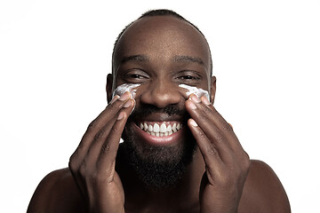 Image showing Young african-american guy applying face cream under his eyes on white background