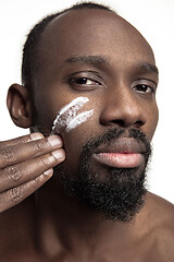 Image showing Young african-american guy applying face cream under his eyes on white background