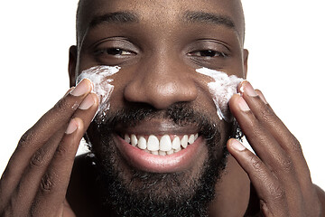 Image showing Young african-american guy applying face cream under his eyes on white background