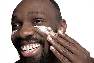 Image showing Young african-american guy applying face cream under his eyes on white background