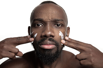 Image showing Young african-american guy applying face cream under his eyes on white background