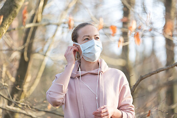 Image showing Portrait of caucasian sporty woman wearing medical protection face mask while walking in park, relaxing and listening to music. Corona virus, or Covid-19, is spreading all over the world