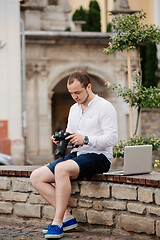 Image showing Young photographer with laptopon stone bench andtaking photo on camera