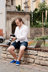 Image showing Young photographer with laptopon stone bench andtaking photo on camera