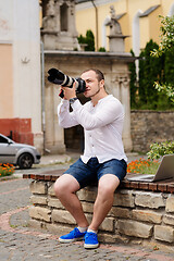 Image showing Young photographer with laptopon stone bench andtaking photo on camera