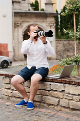 Image showing Young photographer with laptopon stone bench andtaking photo on camera