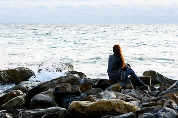 Image showing A girl sits on stony stones by the sea and looks into the distance