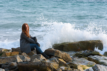 Image showing Girl in autumn windy weather sits by the sea on a rocky shore