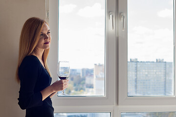 Image showing Beautiful brooding girl with a glass of red wine stands near the window