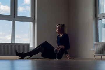 Image showing girl in evening dress and shoes sits on the floor near a large window with wine