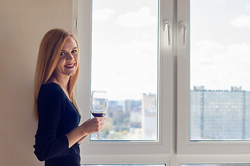 Image showing A girl stands near a window with a glass of wine