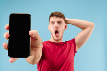 Image showing Young handsome man showing smartphone screen isolated on blue background in shock with a surprise face