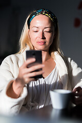 Image showing Beautiful caucasian woman at home, feeling comfortable wearing white bathrobe, taking some time to herself, drinking morning coffee and reading news on mobile phone device in the morning