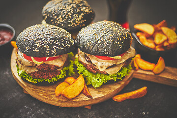 Image showing Homemade delicious black burgers served on wooden cutting board with ketchup, potato wedges