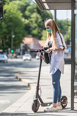 Image showing Casual caucasian teenager commuter waering protective face mask against spreading of corona virus with modern foldable urban electric scooter waiting for metro city bus. Urban mobility concept