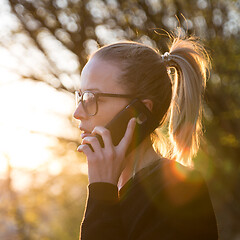 Image showing Backlit rear view of young woman talking on cell phone outdoors in park at sunset. Girl holding mobile phone, using digital device, looking at setting sun