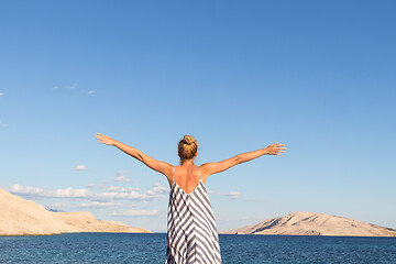 Image showing Happy carefree woman rising arms, wearing beautiful striped summer dress enjoying late afternoon on white pabbled beach on Pag island, Croatia