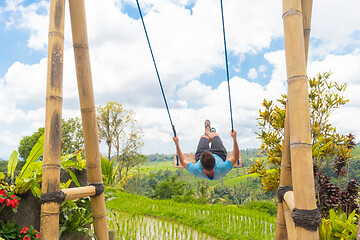 Image showing Happy female traveller swinging on wooden swing, enjoying summer vacation among pristine green rice terraces.