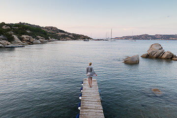 Image showing Beautiful woman in luxury summer dress standing on wooden pier enjoying peaceful seascape at dusk. Vacation, resort and traveling.