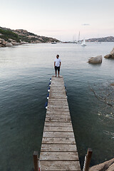 Image showing Man standing on wooden pier enjoying peaceful seascape at dusk. Male tourist stands on a wooden pier in Porto Rafael, Costa Smeralda, Sardinia, Italy