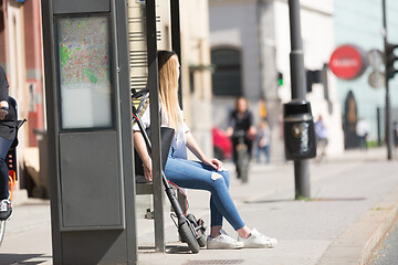 Image showing Casual caucasian teenager commuter with modern foldable urban electric scooter sitting on a bus stop bench waiting for metro city bus. Urban mobility concept