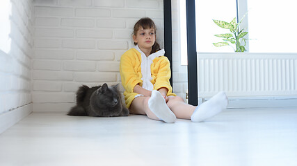 Image showing An upset girl in a yellow bathrobe sits in the corner of the room, a dark gray domestic cat sits nearby