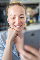 Image showing Young smiling cheerful pleased woman indoors at home kitchen using social media apps on mobile phone for chatting and stying connected with her loved ones. Stay at home, social distancing lifestyle.