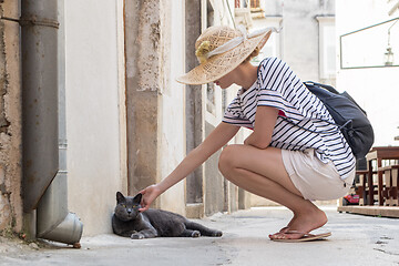 Image showing Female tourist woman wearing big straw hat on summer vacation in ald traditional Mediterranean town, squating on old stone pedestrian street and caressing lazy gray cat lying in front of house