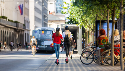 Image showing Rear view of trendy fashinable teenager girls riding public rental electric scooters in urban city environment. New eco-friendly modern public city transport in Ljubljana, Slovenia