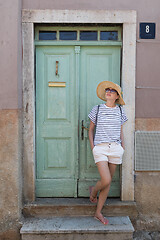 Image showing Beautiful young female tourist woman wearing sun hat, standing and relaxing in shade in front of vinatage wooden door in old Mediterranean town while sightseeing on hot summer day