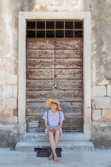 Image showing Beautiful young female tourist woman sitting and resting on vinatage wooden doorstep and textured stone wall at old Mediterranean town, smiling, holding, using smart phone to network on vacationes