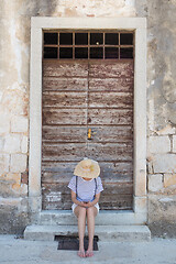 Image showing Beautiful young female tourist woman sitting and resting on vinatage wooden doorstep and textured stone wall at old Mediterranean town, smiling, holding, using smart phone to network on vacationes