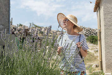 Image showing Beautiful blonde young female traveler wearing straw sun hat enjoying summer on Mediterranean cost strolling among lavander flowers on traditional costal village garden