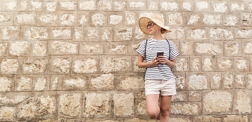 Image showing Beautiful young female tourist woman standing in front of old textured stone wall at old Mediterranean town, smiling, holding, smart phone to network on vacationes. Copy space