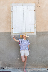 Image showing Beautiful young female tourist woman wearing big straw hat, taking self portrait selfie, standing in front of white vinatage wooden window and textured stone wall at old Mediterranean town